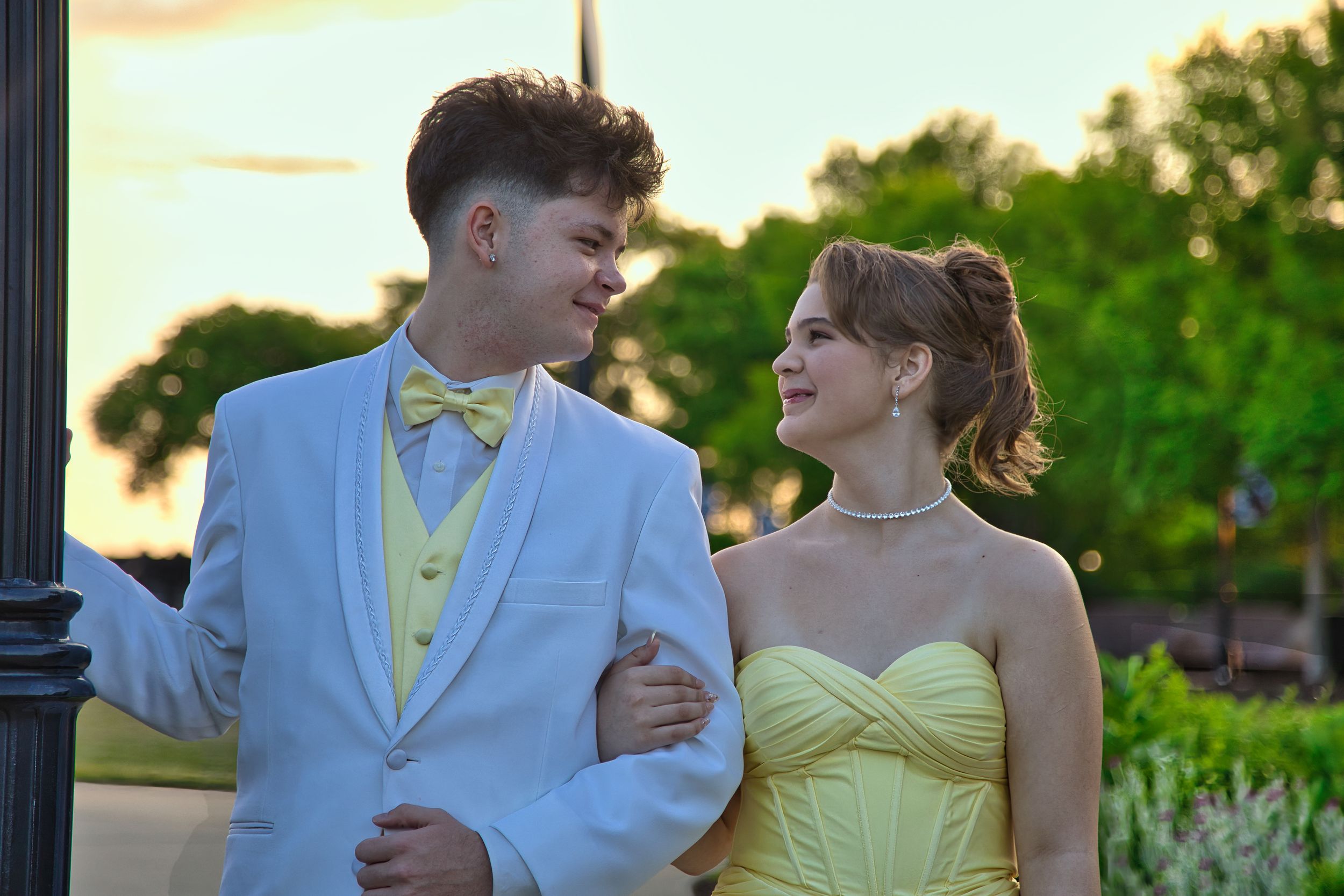 Smiling prom couple in matching yellow and white formal attire posing together near lamppost.