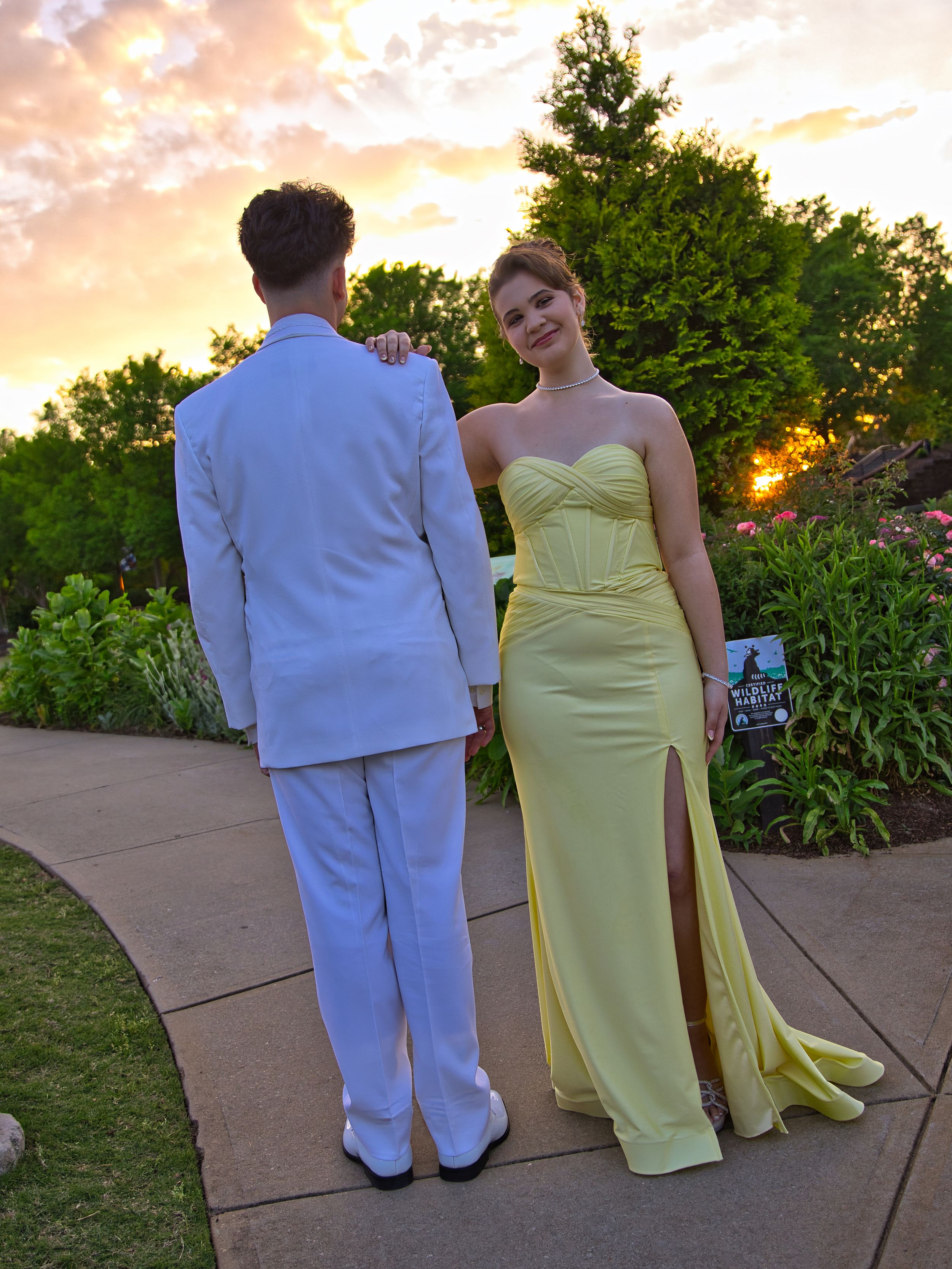 Prom couple walking on garden path at sunset in white tuxedo and yellow mermaid gown.