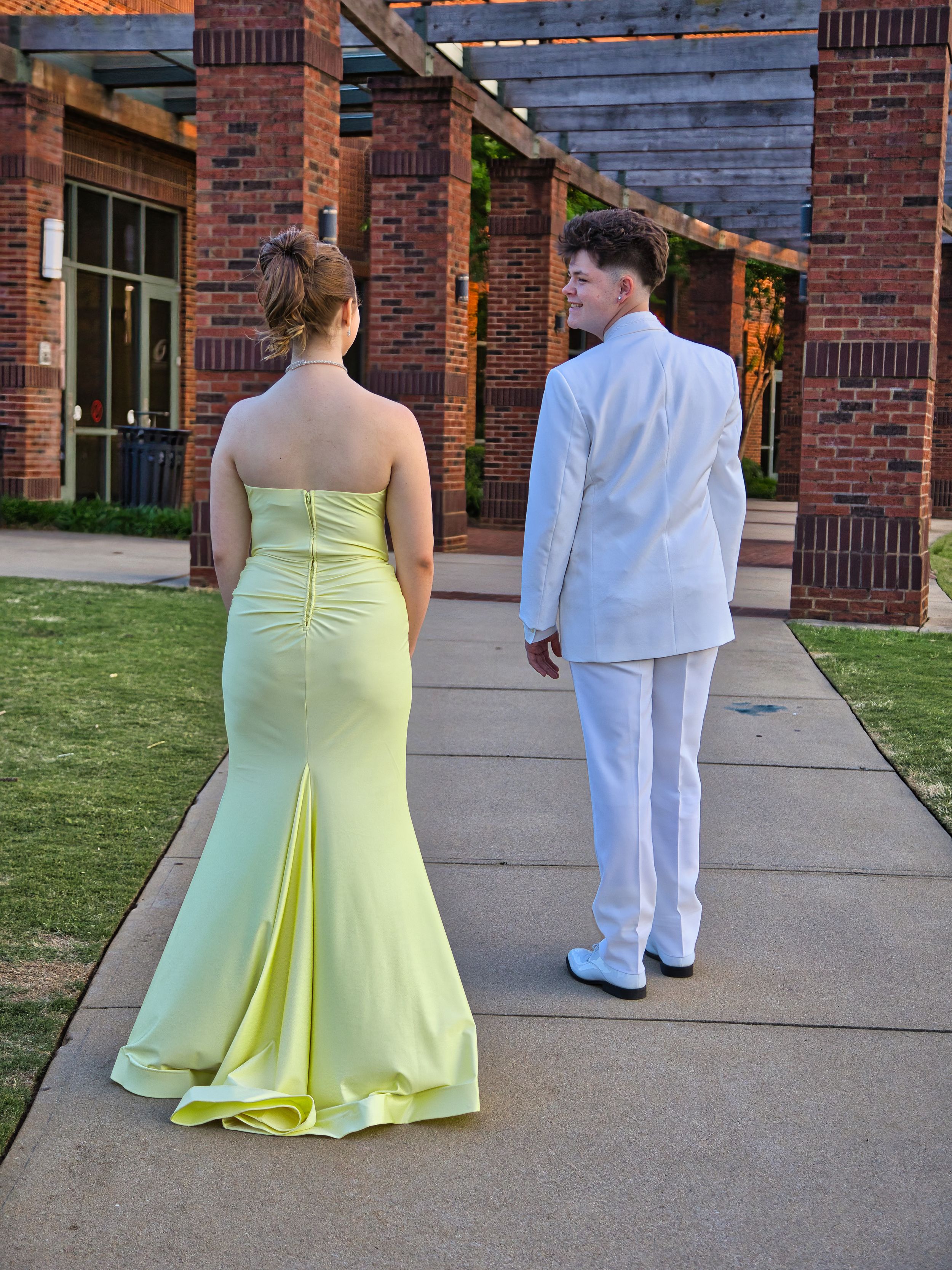 Prom couple walking on sidewalk near brick building, girl in yellow gown and boy in white tux.