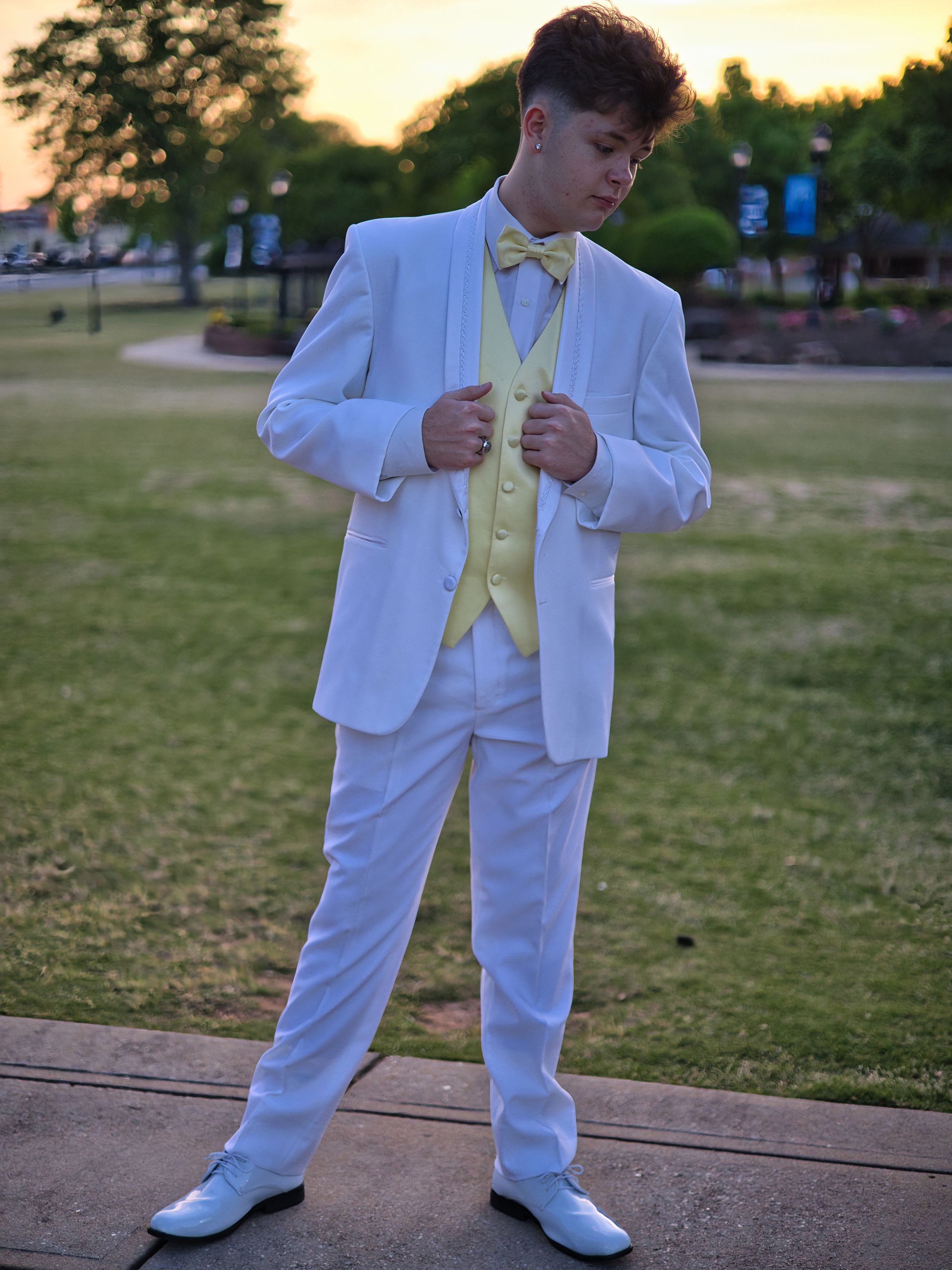 Teen boy in white formal suit with yellow vest and bow tie adjusting jacket at sunset outdoors.