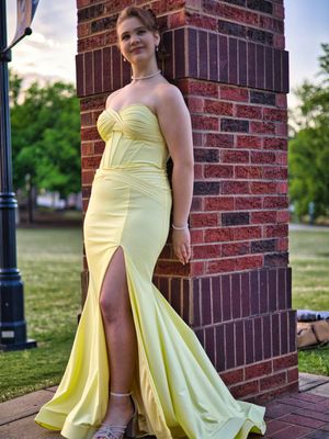Young woman in strapless yellow mermaid prom dress with slit posing against brick column at dusk.