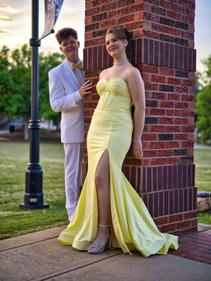 Prom couple posing together, boy in white tuxedo and girl in yellow strapless mermaid gown outdoors.