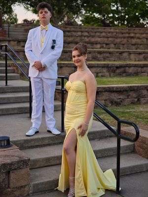 Prom couple standing on outdoor steps, boy in white suit and girl in elegant yellow formal gown.