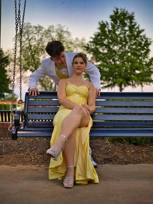 Prom couple on outdoor swing bench, girl in yellow gown seated with boy standing behind her.
