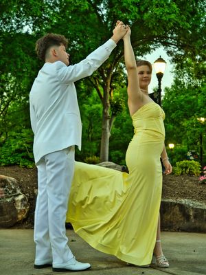 Prom couple dancing outdoors at dusk, boy in white suit spinning girl in flowing yellow gown.