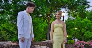 Prom couple in white suit and yellow strapless gown standing together in lush garden setting.