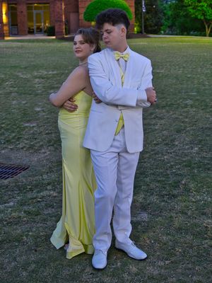 Prom couple posing back-to-back outdoors in yellow gown and white suit with yellow bow tie.