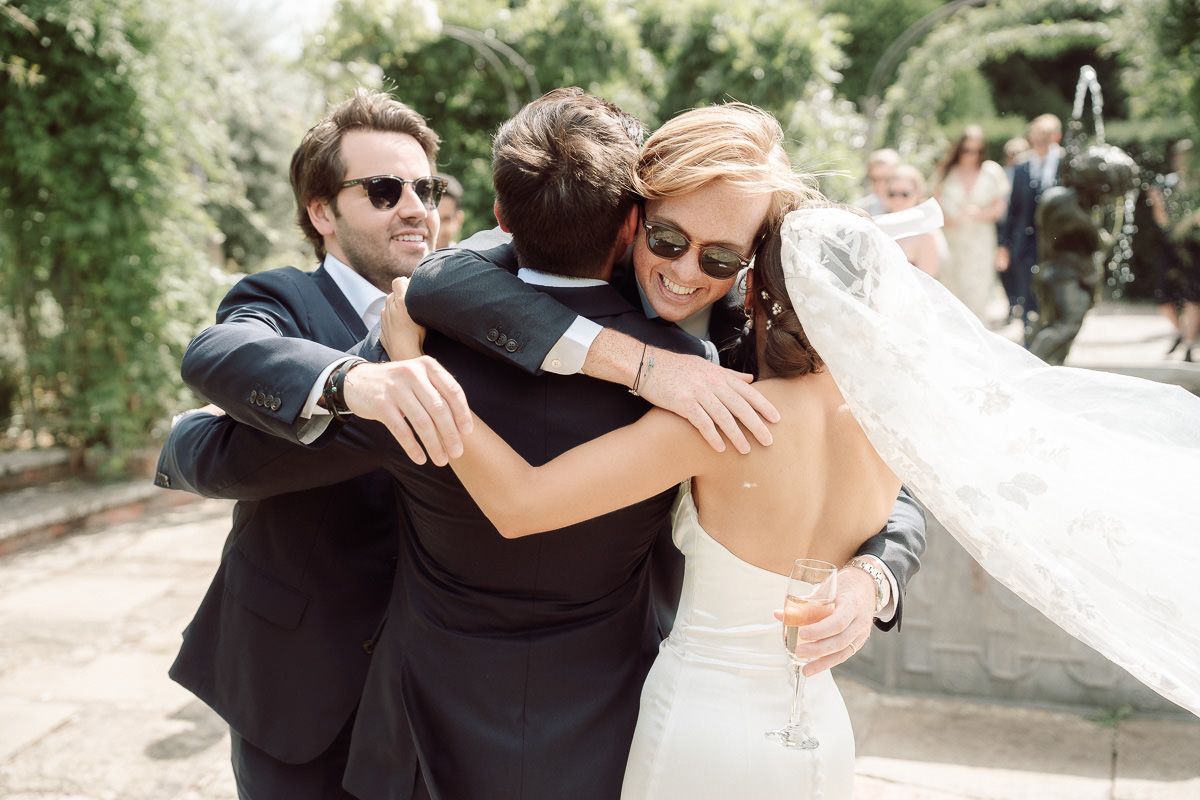 Bride and groom share a group hug with a guest in sunglasses at an outdoor wedding celebration.
