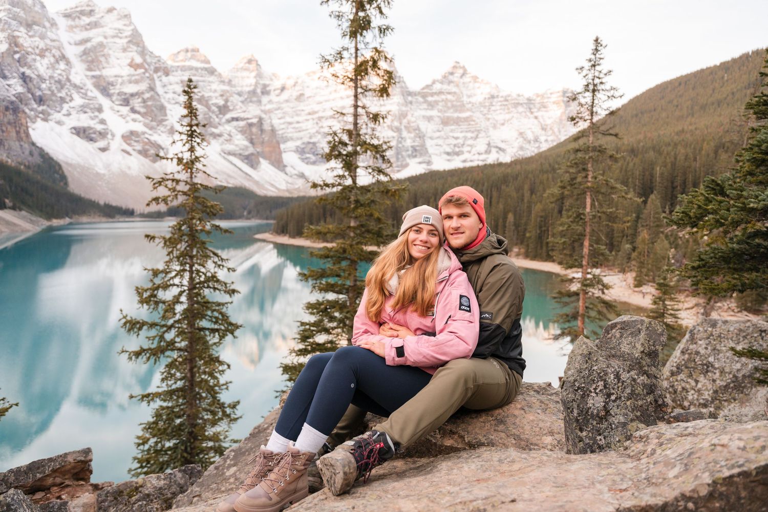 Couple sitting on rocks at Moraine Lake, Banff, with turquoise water and snow-capped mountains behind them.