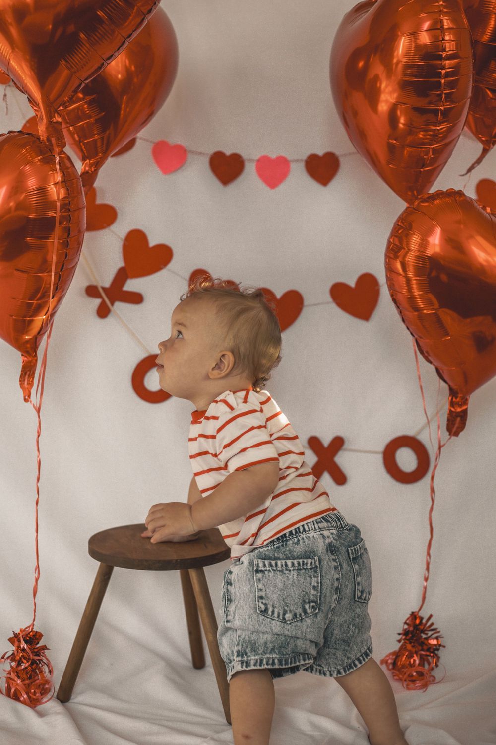 Toddler in striped shirt and denim shorts stands by wooden stool amid red heart balloons and XOXO Valentine's Day decor.