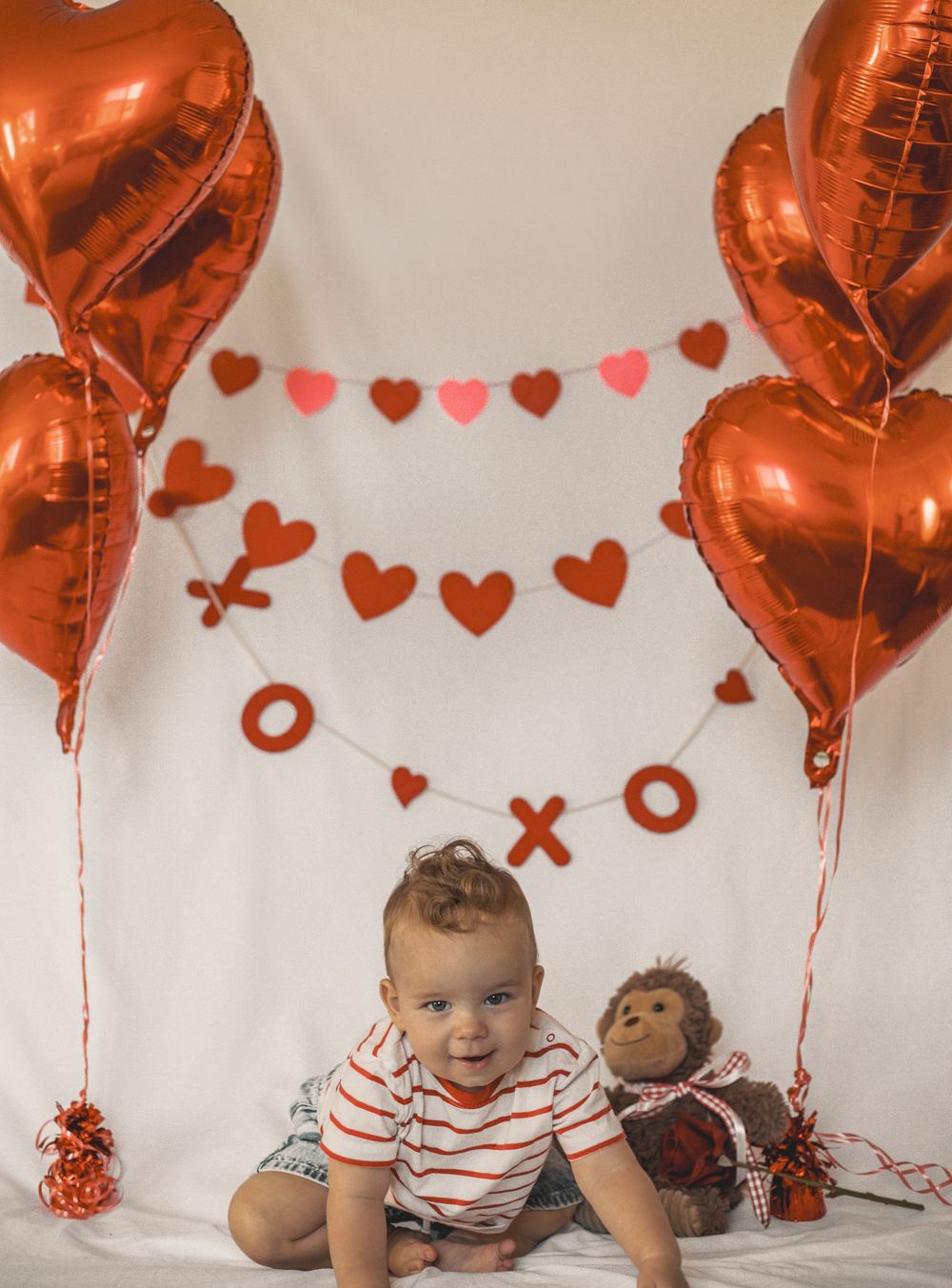 Baby boy in striped shirt surrounded by red heart balloons and Valentine's Day decorations on white backdrop.