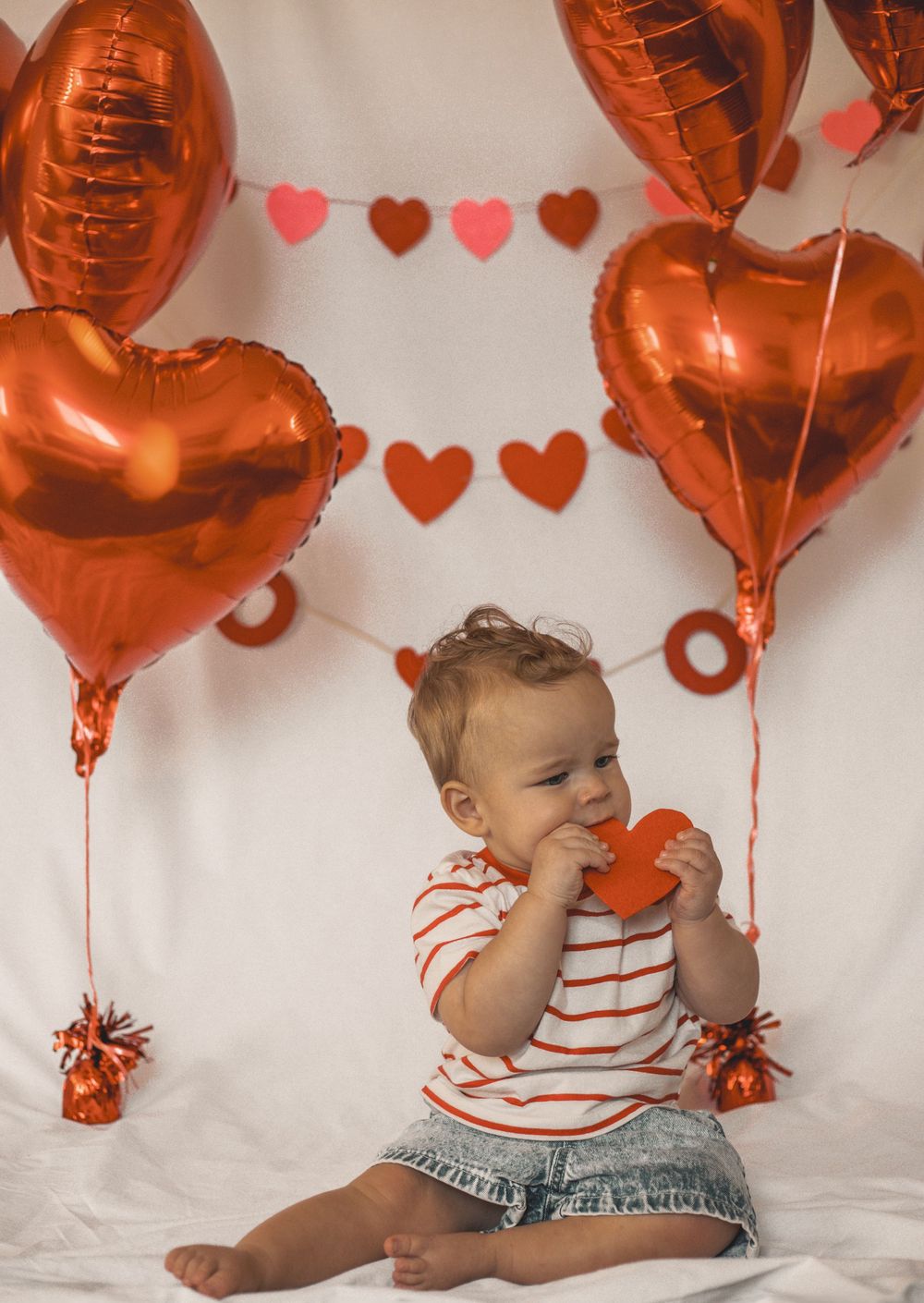 A baby sits surrounded by red heart balloons and heart garland, holding a red heart cutout in a Valentine's Day photo.