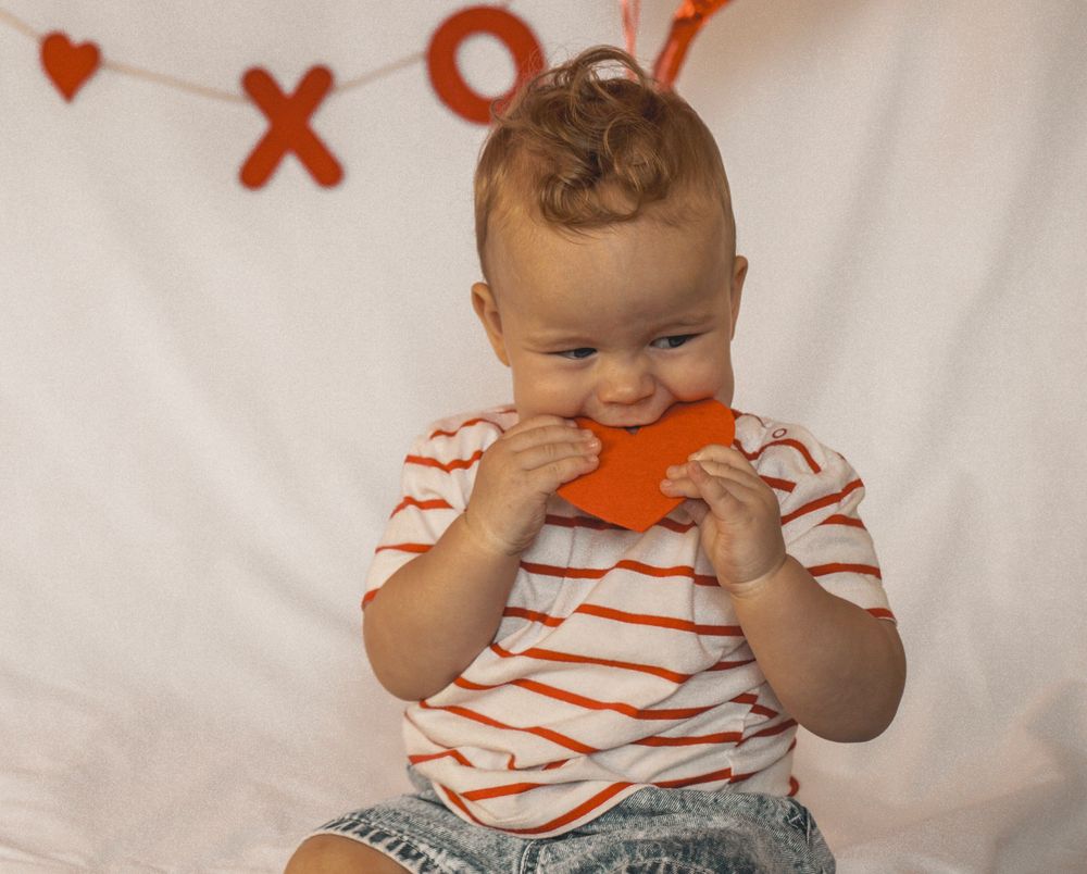 Baby in striped shirt chewing orange star toy with XO Valentine's Day decorations in background.