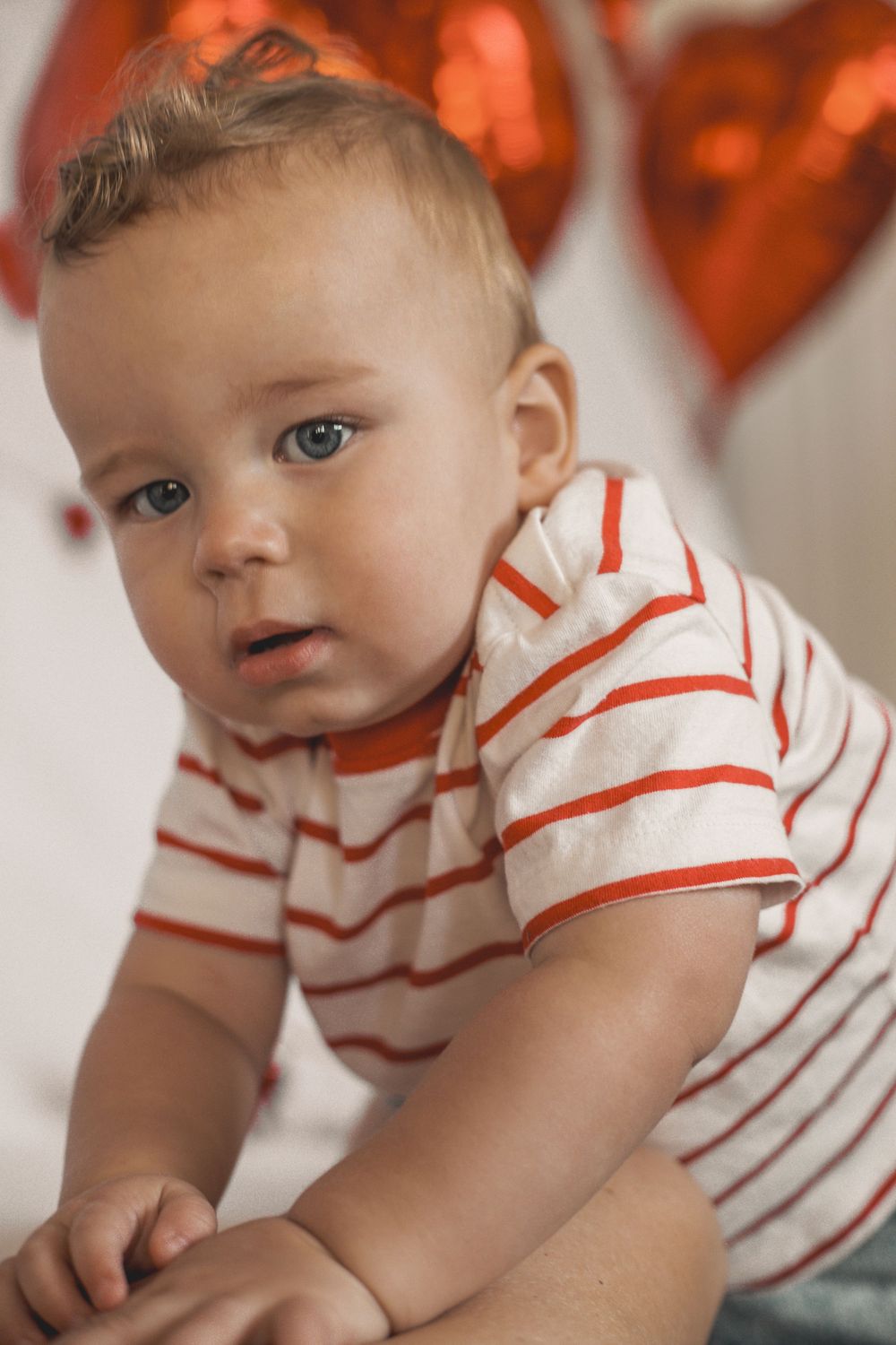 Adorable baby boy in red striped shirt sitting near heart-shaped red balloons, looking curiously at camera.