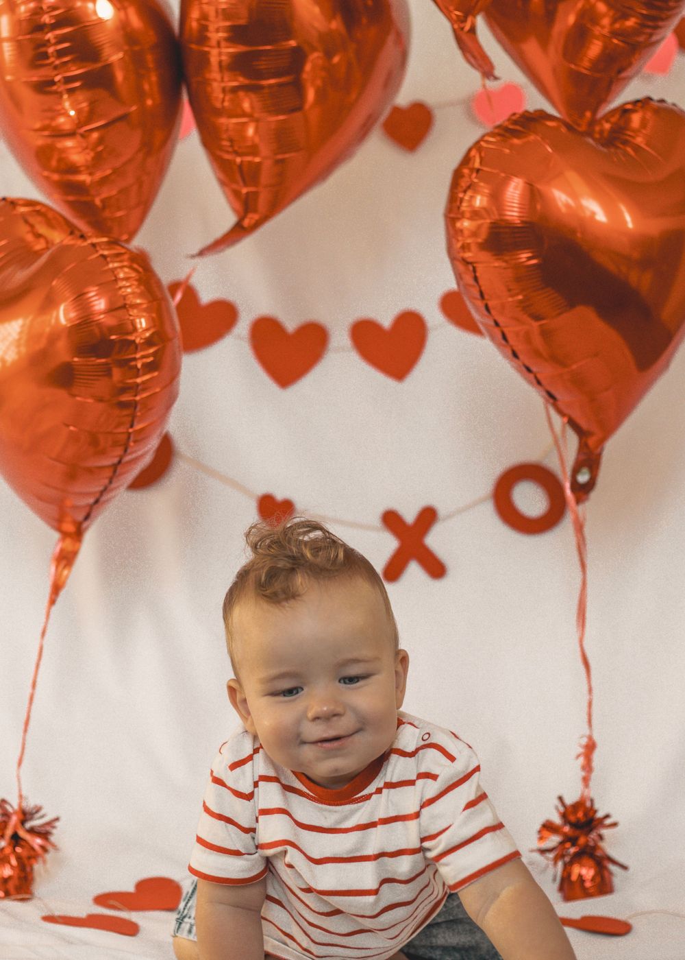 Smiling baby in striped shirt sits before red heart balloons and Valentine decorations on white backdrop.