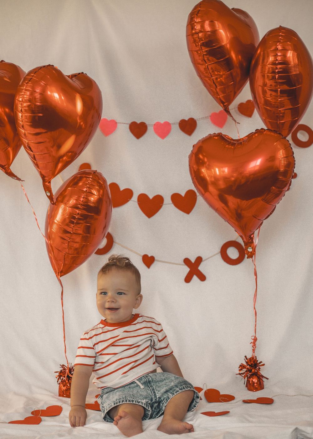 Happy baby sitting among red heart balloons and Valentine decorations on white backdrop.