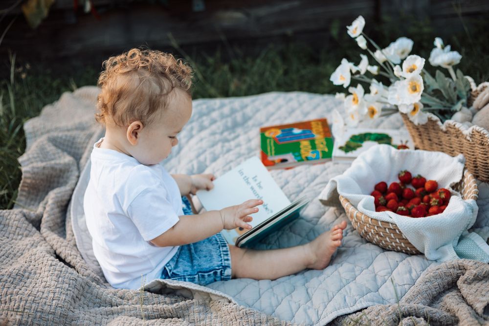 A curious toddler sits on a blanket outdoors, reading a book near a basket of fresh strawberries and white flowers.