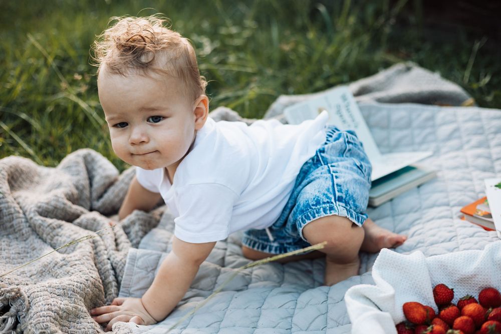 A cute baby boy in a white shirt and denim shorts crawling on a blanket outdoors near fresh strawberries.