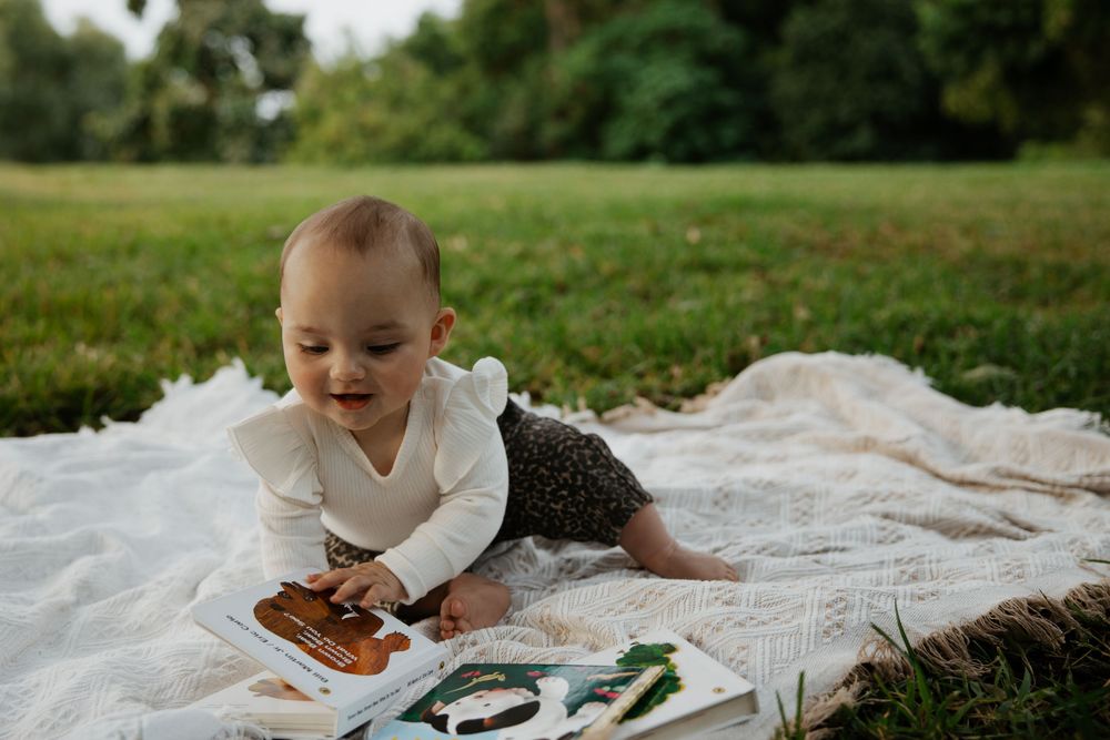 Baby lying on a blanket in a park, happily exploring a colorful picture book outdoors.