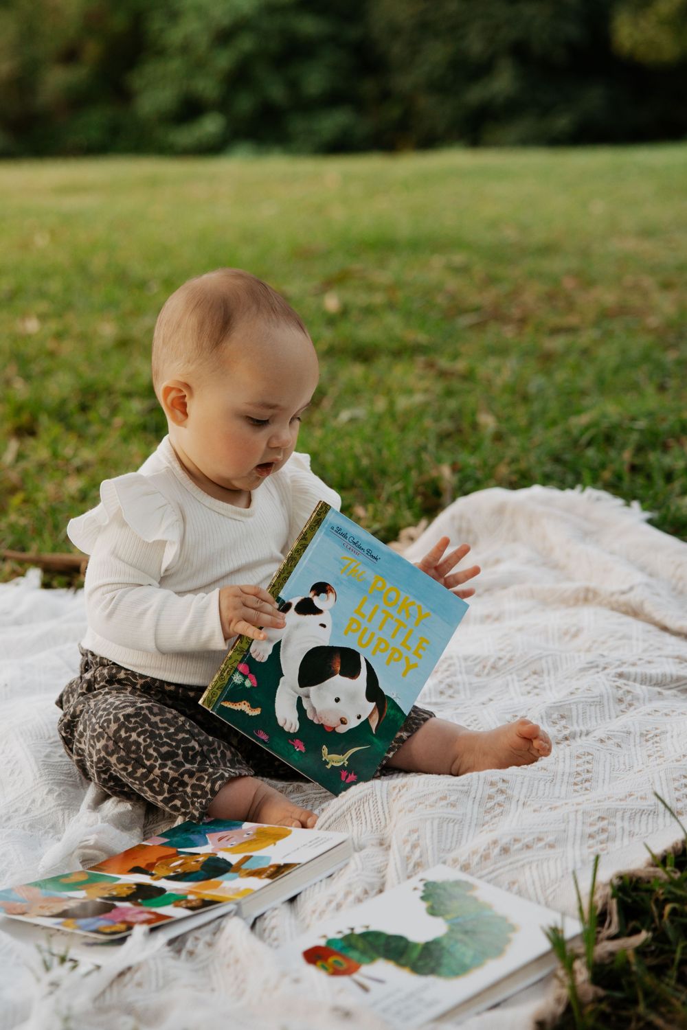 A baby girl sits on a white blanket in a park, reading a Little Golden Book surrounded by colorful picture books.