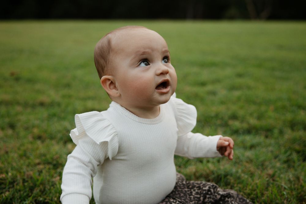 Adorable baby in white ruffle onesie sitting on green grass, looking up with curious expression outdoors.