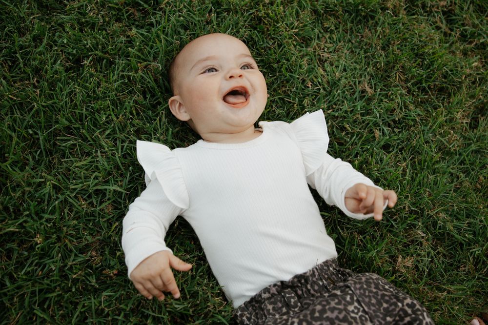 A happy baby in a white ruffle shirt laughs while lying on lush green grass outdoors.