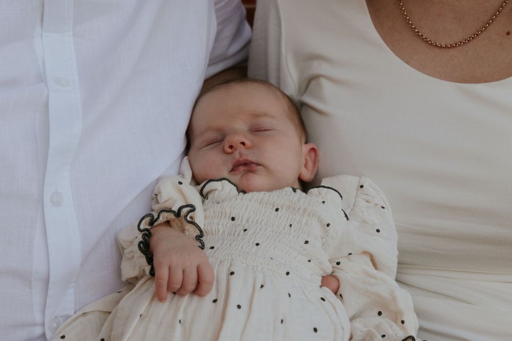 Sleeping newborn baby in white smocked polka dot gown cradled between parents wearing white clothing.