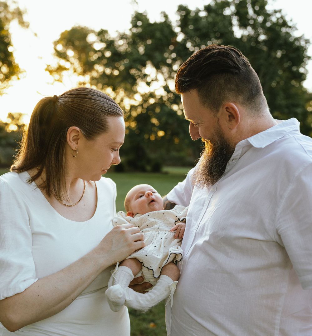 A couple in white clothing lovingly gazes at their newborn baby outdoors in golden hour sunlight.