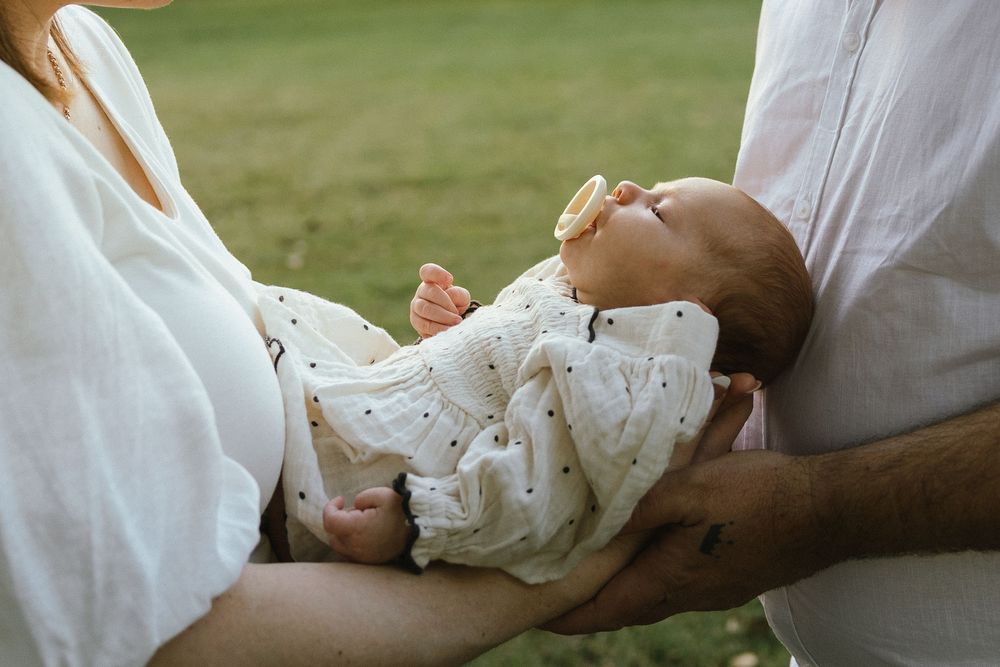 Parents cradling a sleeping newborn baby wrapped in a white muslin swaddle with a pacifier outdoors.