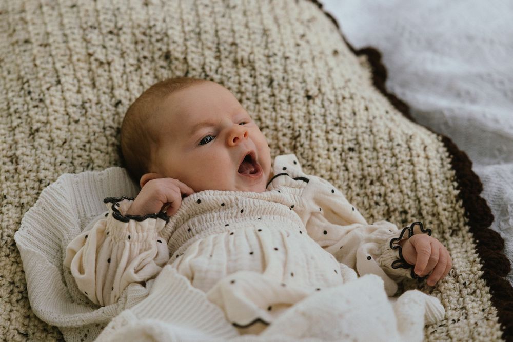 Newborn baby yawning while lying on a textured cream knit blanket, dressed in a white dotted romper.