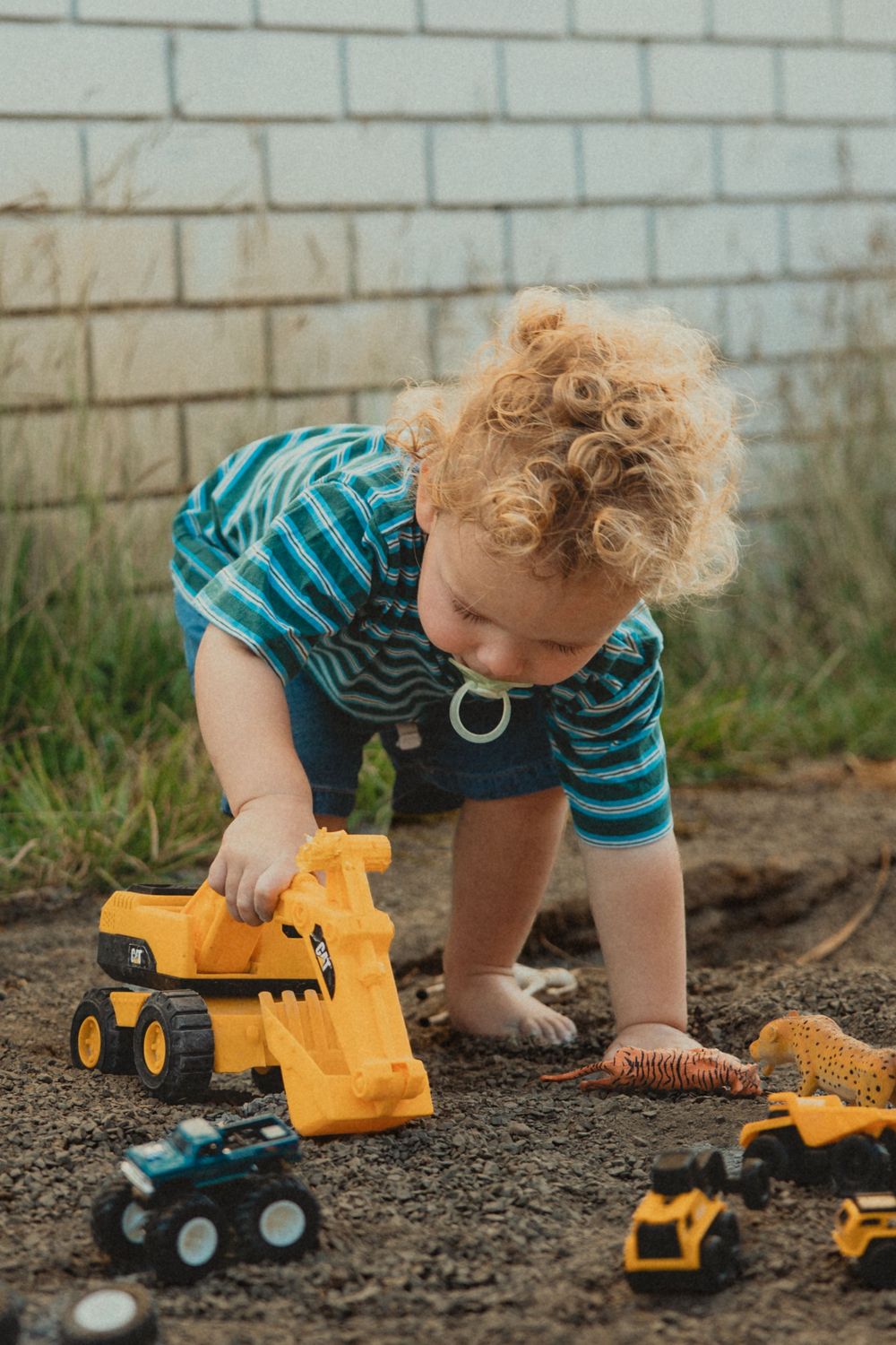 Curly-haired toddler in a striped shirt plays with yellow toy construction trucks and diggers in the dirt beside a white brick wall.