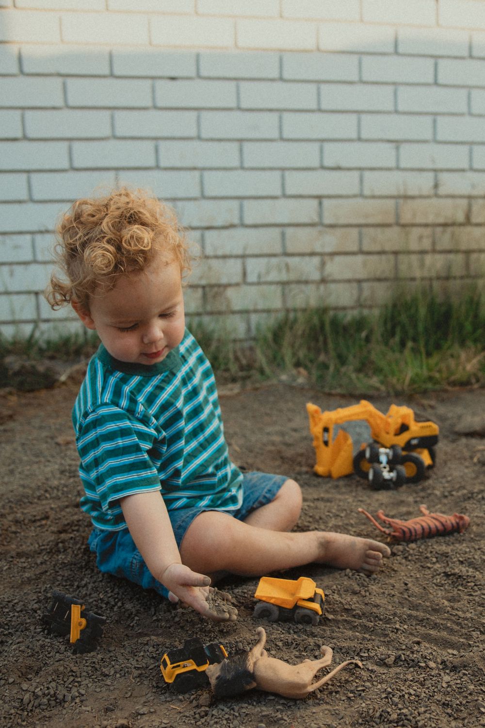 Curly-haired toddler in a striped teal shirt sits in the dirt playing with yellow toy excavators and dinosaur figures by a white brick wall.
