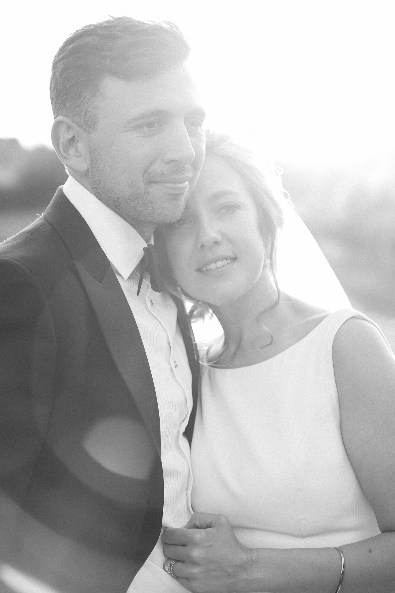 Black and white wedding portrait of a bride and groom embracing in soft backlight, smiling tenderly together at Lightfoot & Wolfville, NS