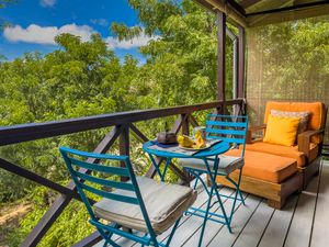 Screened balcony with blue bistro chairs and table, orange lounge chair, overlooking lush green tropical treetops.