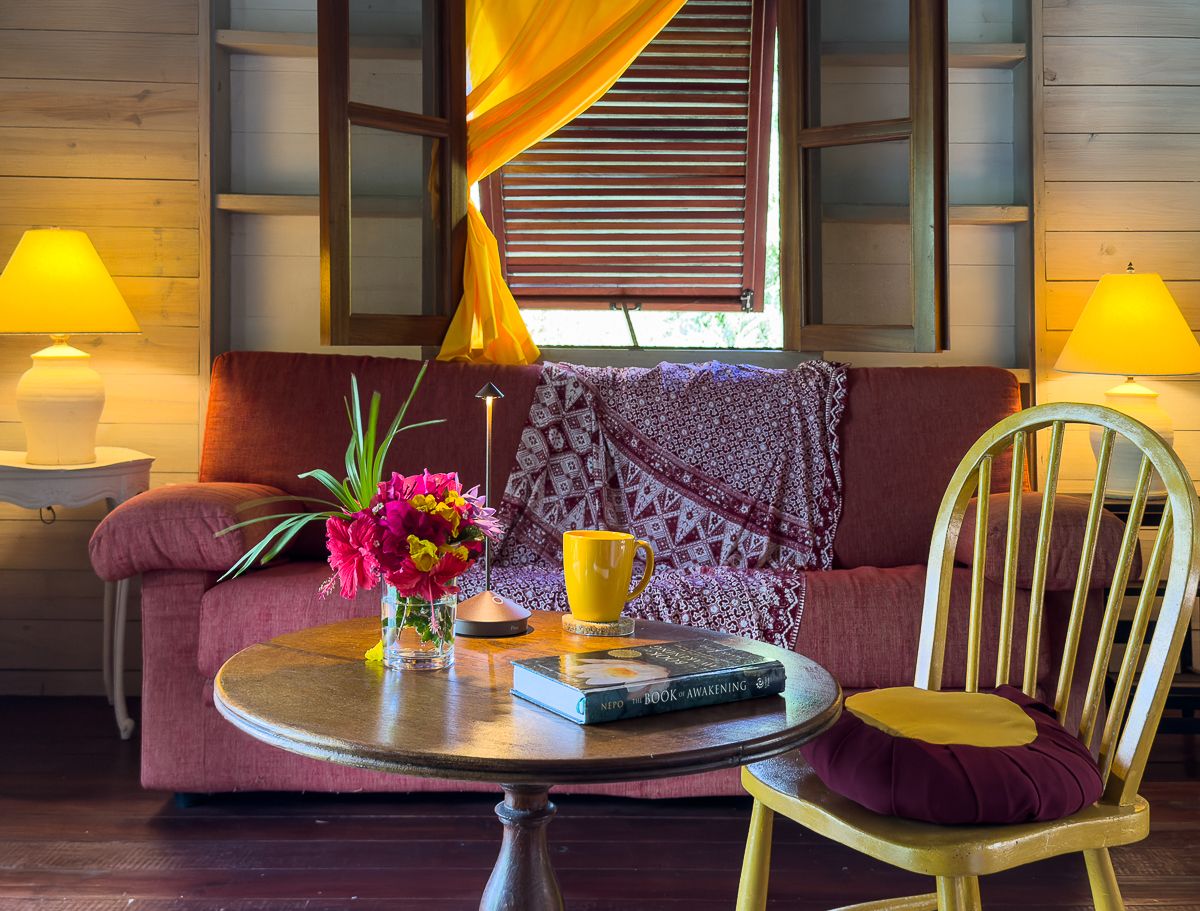 Cozy tropical living room with red sofa, yellow lamps, wooden walls, and a round table with fresh flowers.