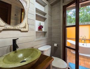 Rustic bathroom with green vessel sink, ornate mirror, built-in shelves, toilet, and outdoor clawfoot tub on wooden deck.