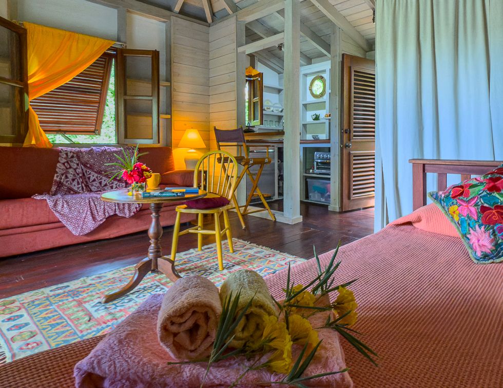 Cozy tropical cottage interior with pink sofas, colorful decor, yellow chairs, and canopy bed in warm Caribbean-style room.
