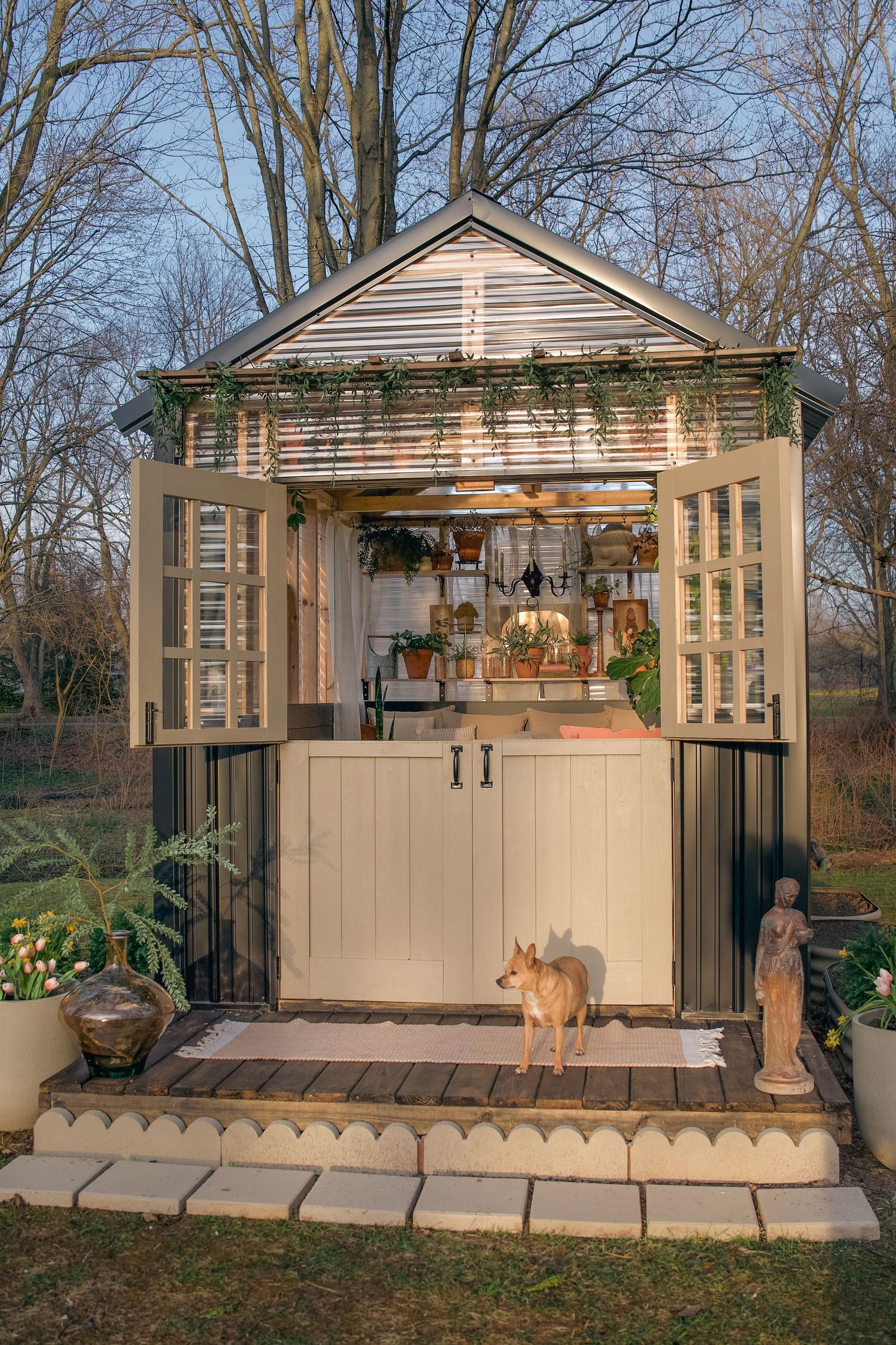 Charming garden shed with open shuttered windows, hanging plants, a dog on the porch, and a stone statue at dusk.