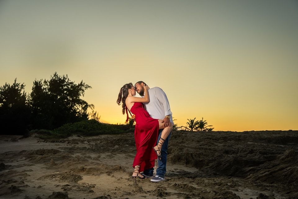 Couple sharing a romantic kiss at sunset on sandy dunes, woman in red dress, golden sky backdrop.