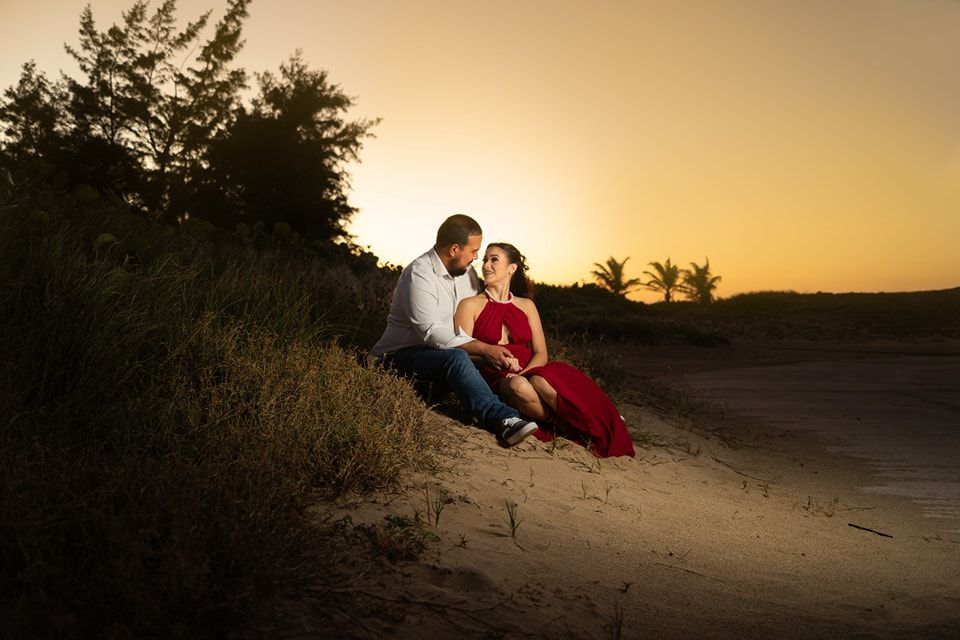 Couple sitting together on sandy beach dunes at golden sunset, woman in red dress, romantic engagement photo.