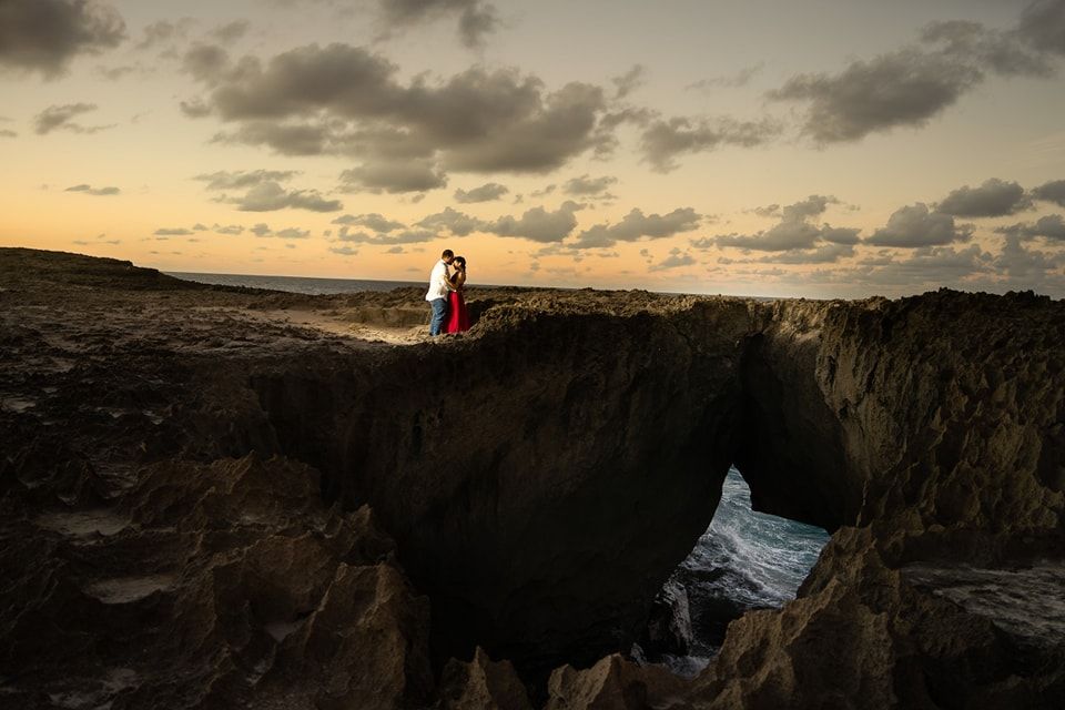 Couple embraces on dramatic coastal cliffs at sunset, with ocean waves crashing below and golden clouds above.