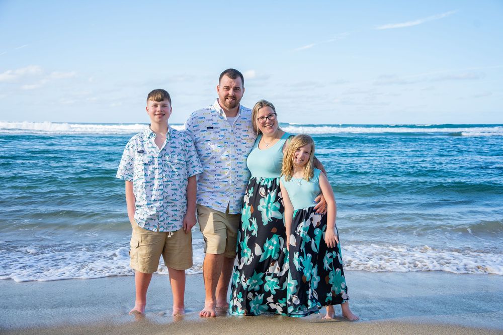 Family of four smiling together on a sunny beach with turquoise waves, wearing coordinated outfits in teal and white.