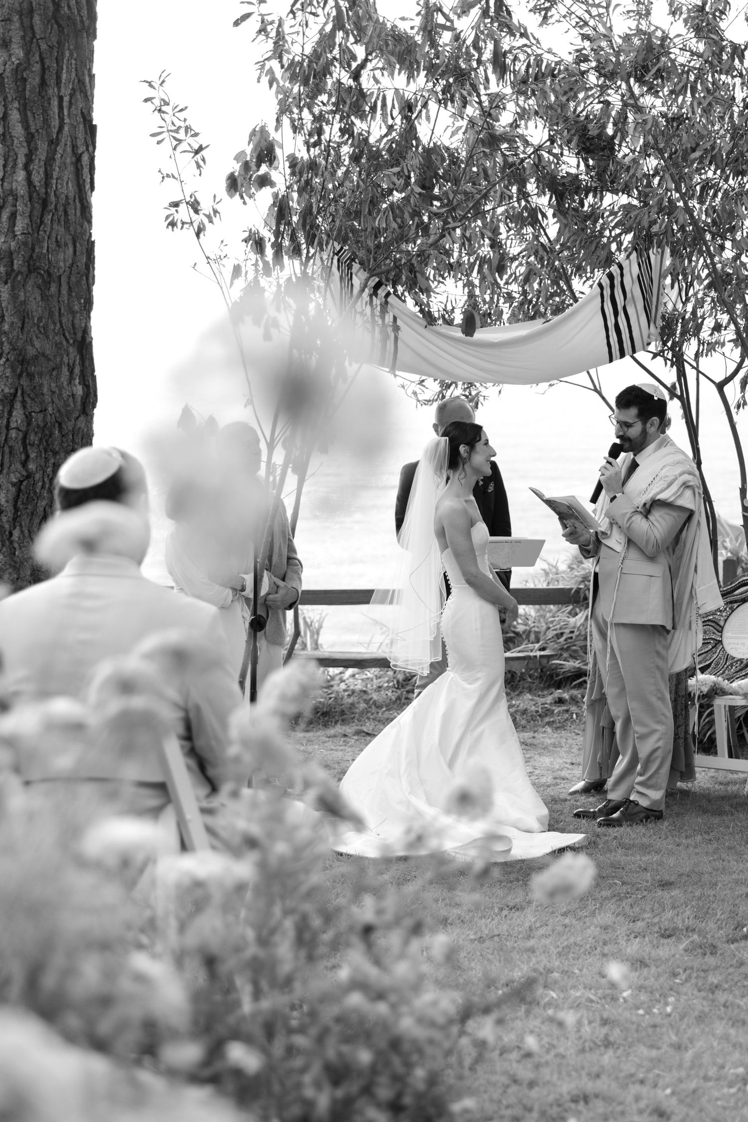 Black and white outdoor Jewish wedding ceremony under chuppah near water with officiant reading.