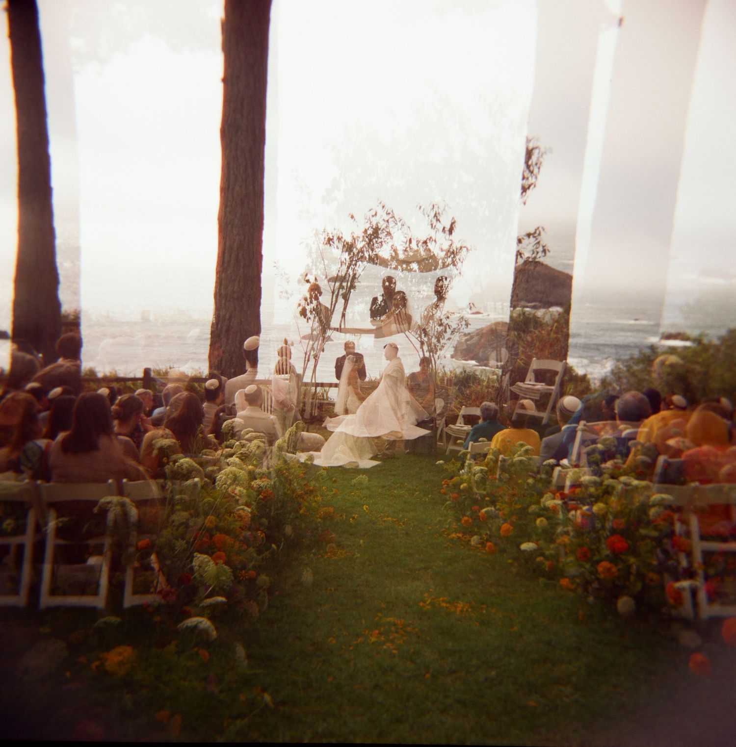 Outdoor wedding ceremony with couple at altar, ocean view backdrop, floral aisle, and seated guests on lush lawn.