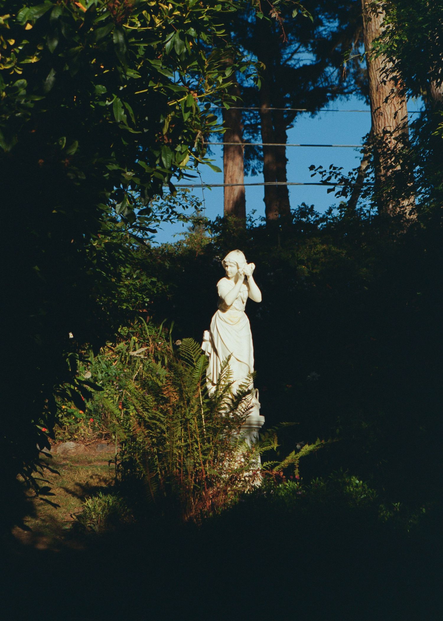 White classical garden statue of a woman surrounded by lush green foliage in dappled sunlight.