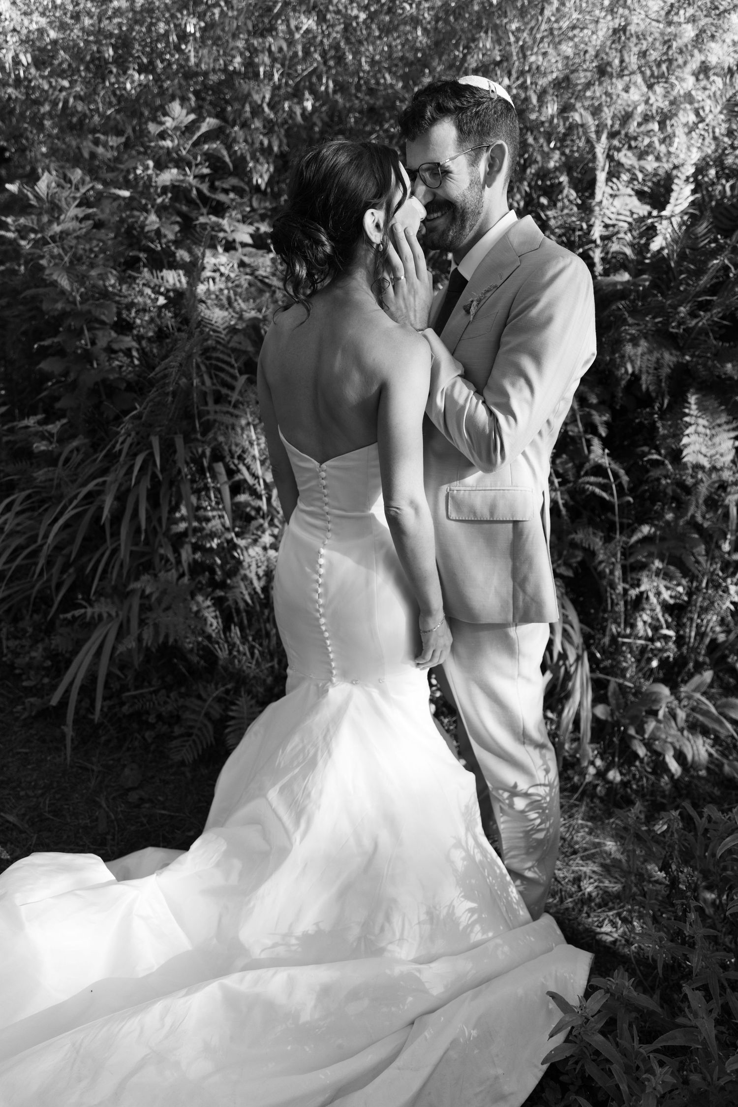 Groom tenderly cups brides face in black and white romantic wedding portrait surrounded by greenery.