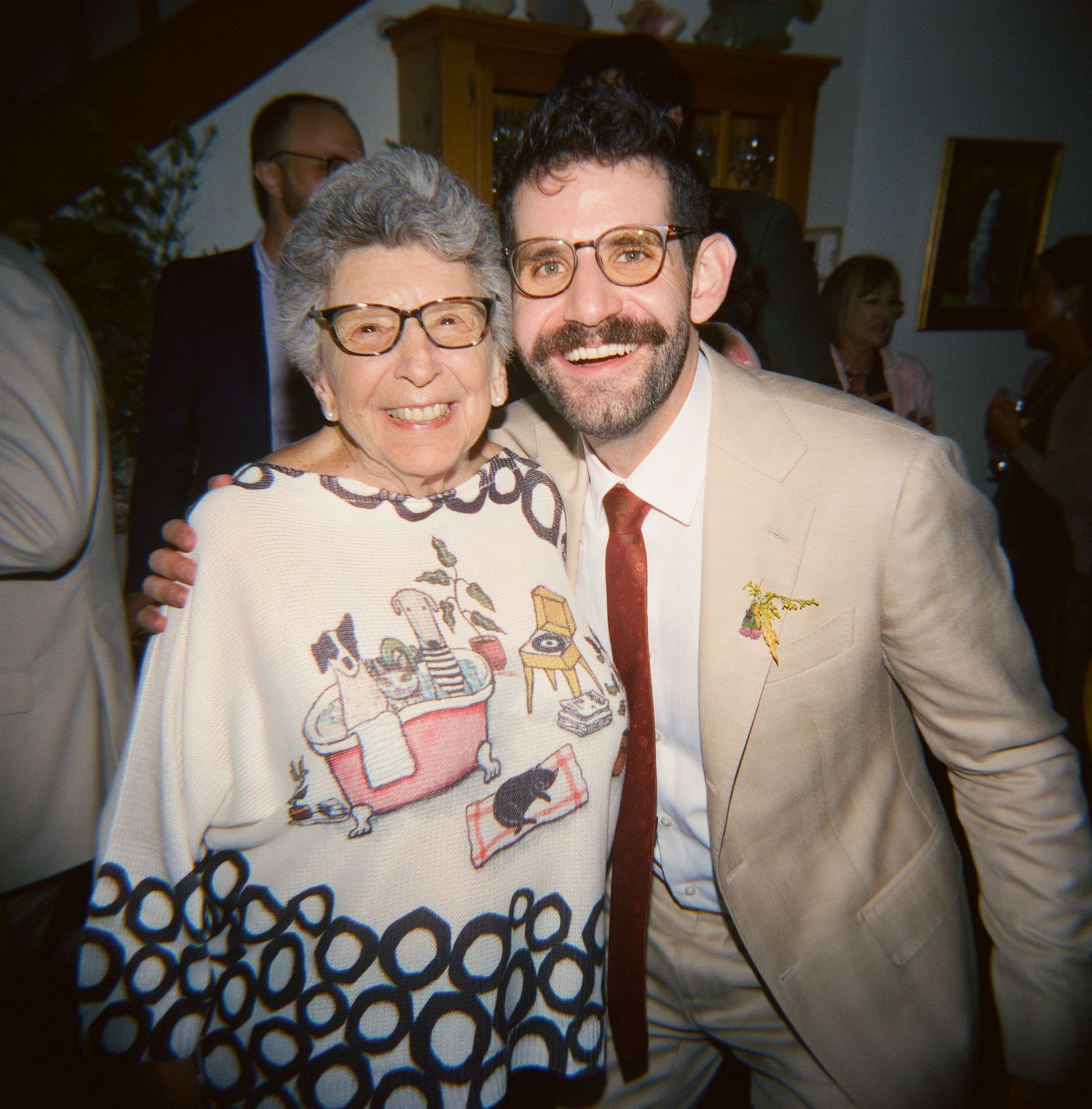 Elderly woman and bearded man in cream suit smiling together at an indoor wedding reception.