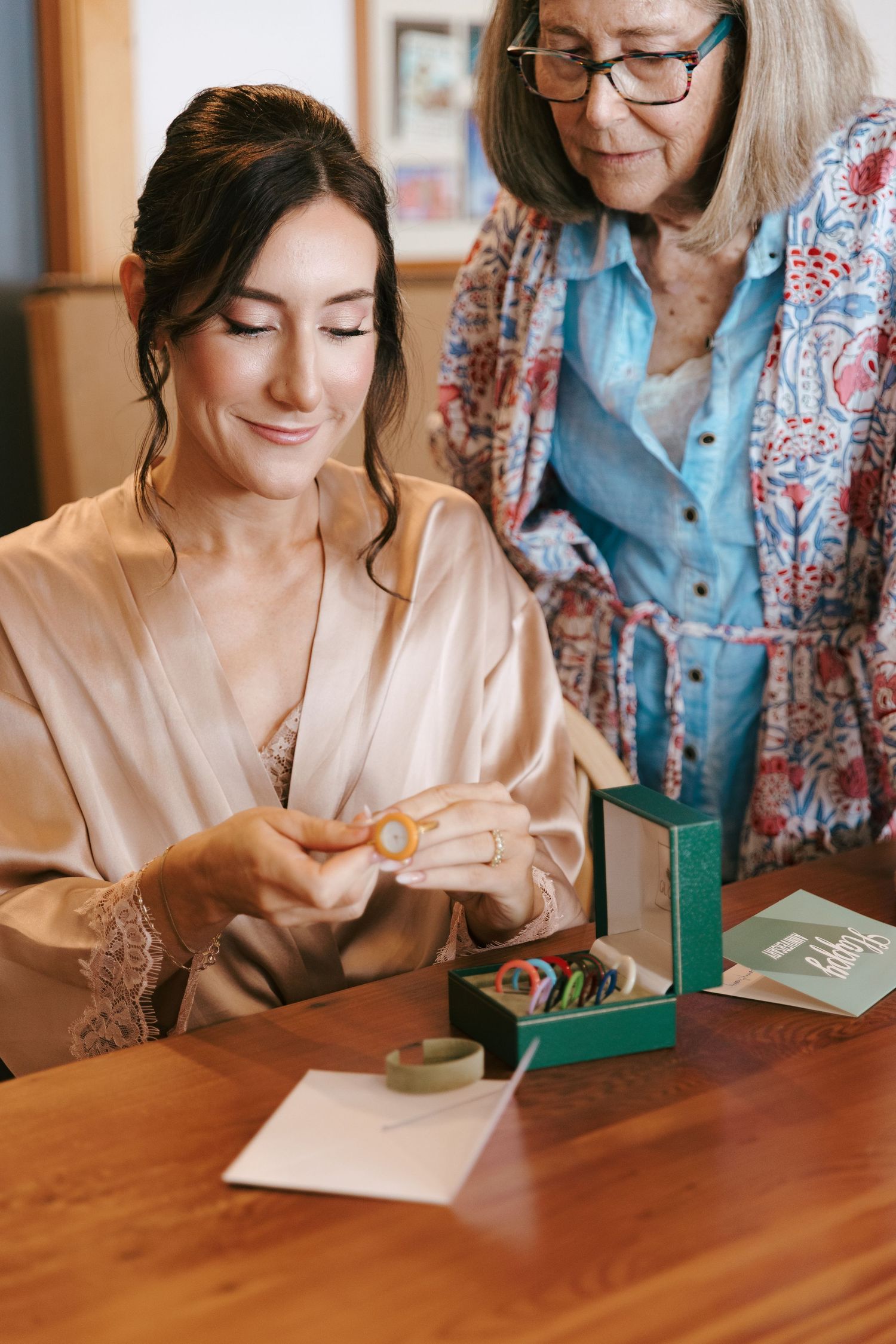 Bride in silk robe opens jewelry gift box with older woman at wooden table.