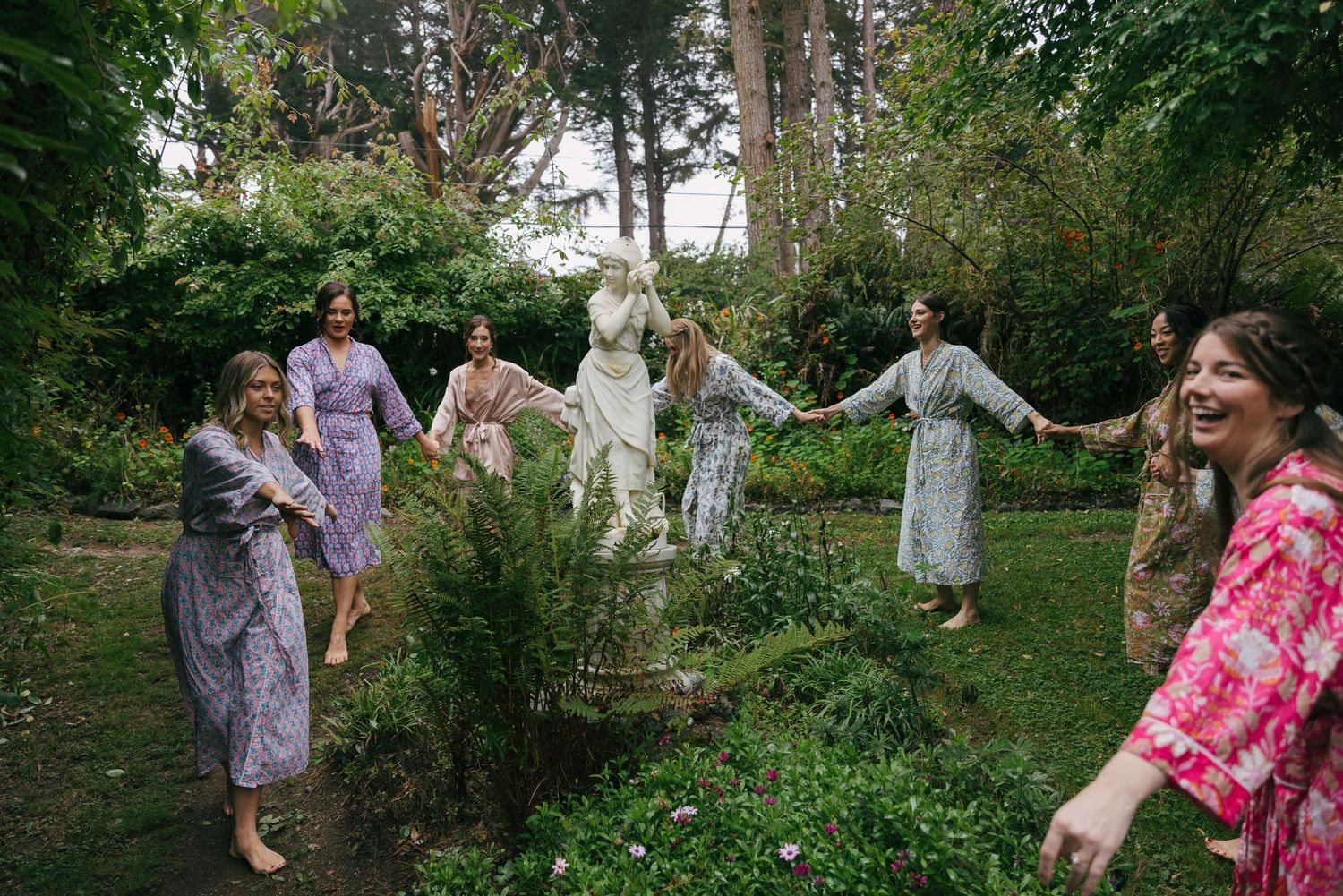 Bridesmaids in floral robes dancing around a garden statue amid lush greenery before the wedding.