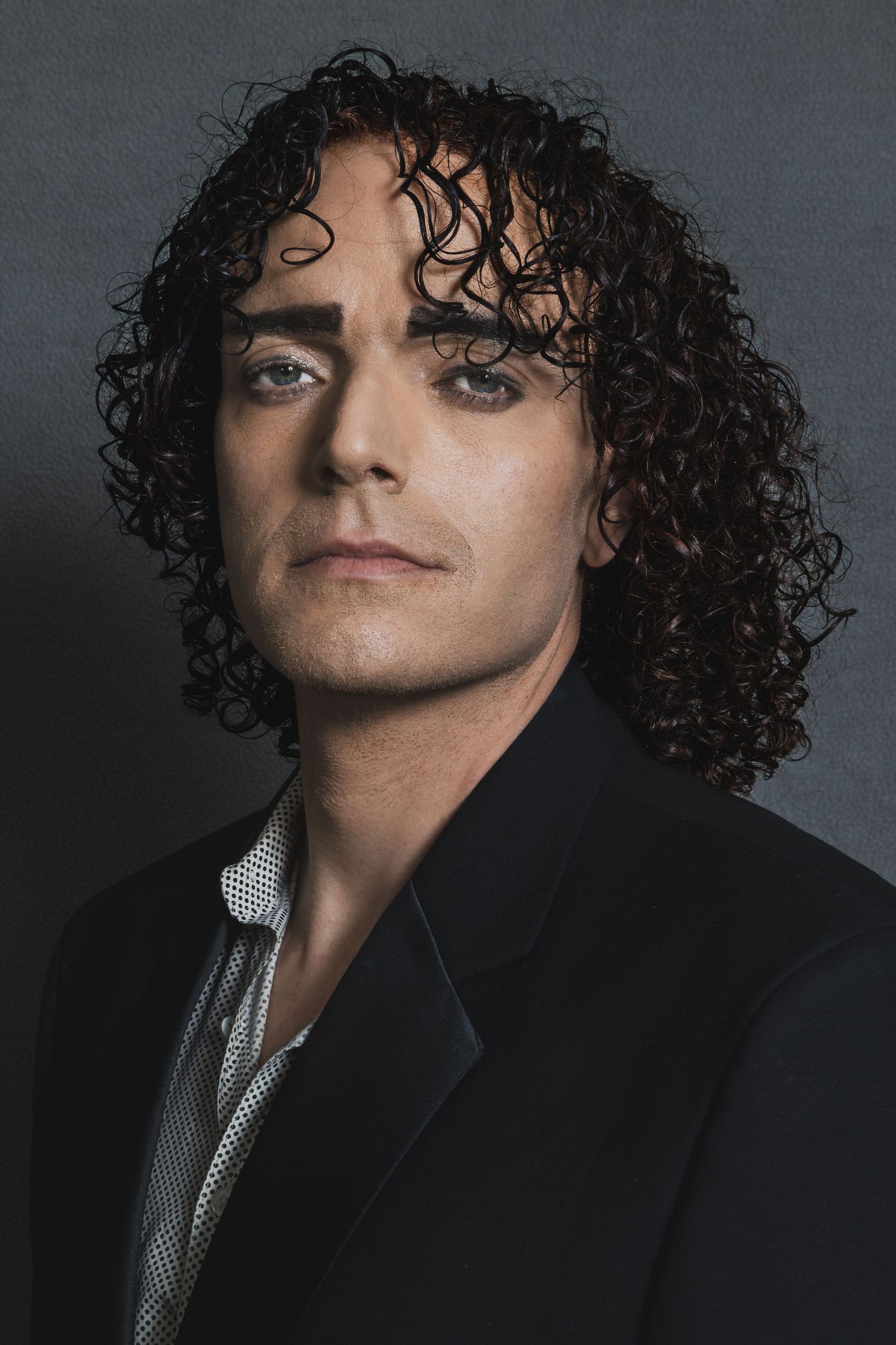 Young man with long dark curly hair wearing a black suit and silver tie poses against a gray background.